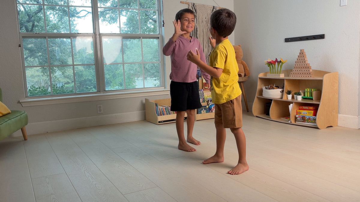 Two boys high-five in a tidy playroom after resetting a Dannico front-facing bookshelf and Joy Shelf toy shelf