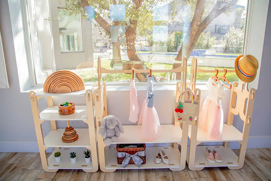 Custom wooden children's wardrobe and storage unit by Dannico Woodworks, featuring a playful and functional design with shelves and hanging space. Montessori-style rainbow stacker, plush toys, and children's clothing tastefully displayed against a serene backdrop with a view of a tree and pond, perfect for an organized and stylish nursery or playroom