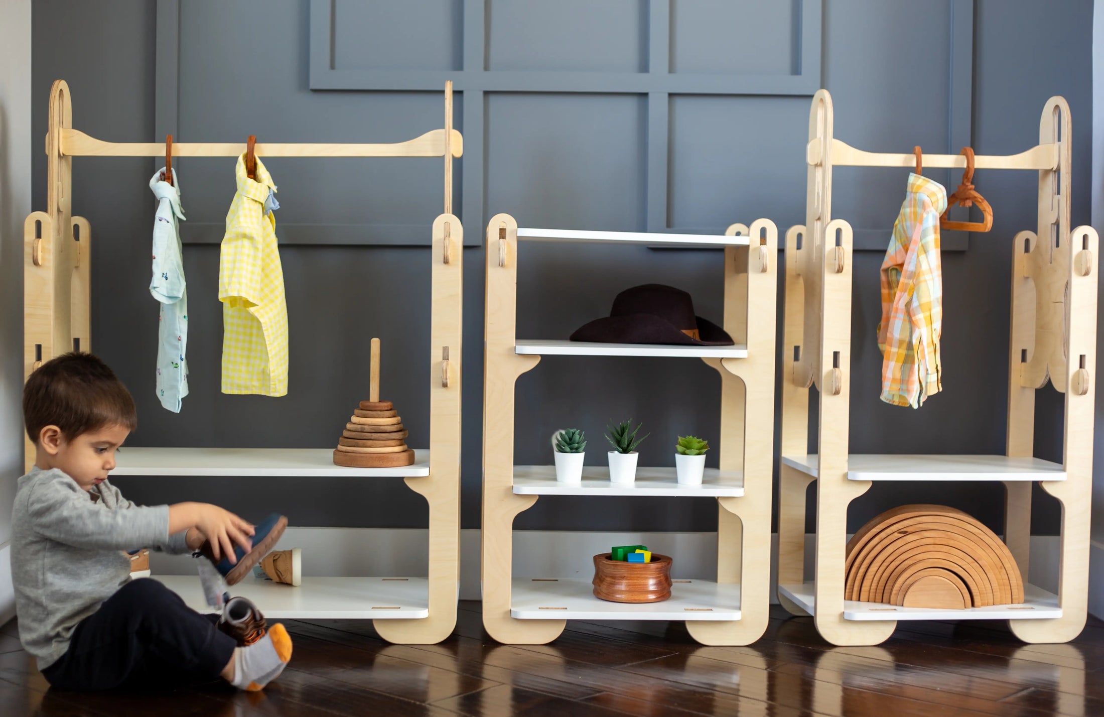 Alt text: "A cozy corner of a children's playroom, with a young child playing on a dark wooden floor. The room features elegant, natural wooden furniture: two clothing racks with whimsical cut-outs, one displaying a yellow dress and another a multicolored shirt. A shelving unit stands in the middle, adorned with a stylish dark hat, a stack of wooden rings, and several potted succulents. A wooden rainbow toy rests on the lower shelf, adding a touch of playful charm to the scene.