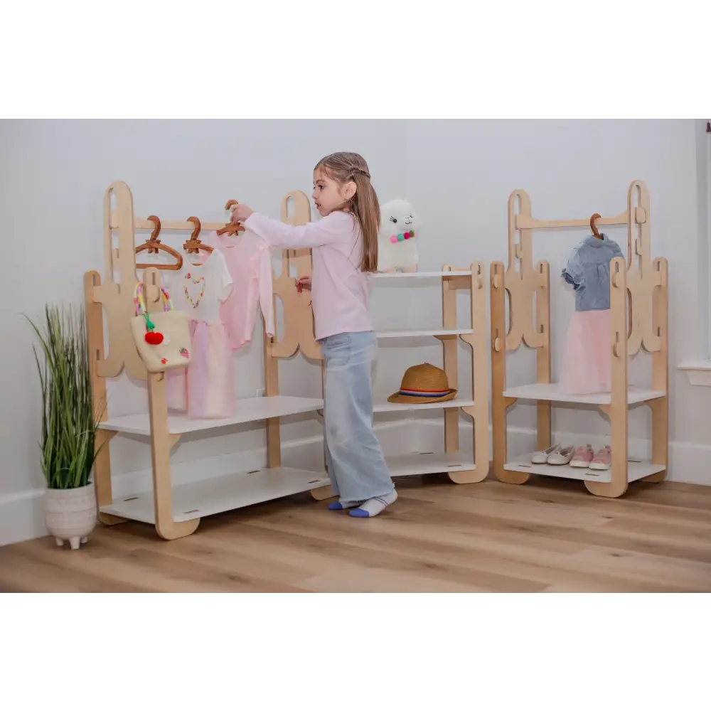 Little girl carefully hanging dresses on a beautifully crafted wooden clothes rack, with a playful stuffed animal on the top shelf and various children's shoes on the bottom, perfect for a tidy and charming child's room