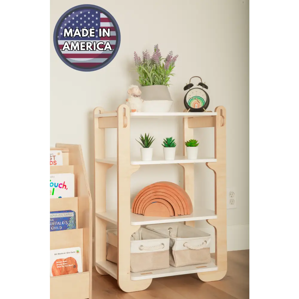 A warm and neatly organized wooden shelf in a child's room, labeled 'Made in America'. The top shelf hosts a lavender plant in a white pot alongside a vintage black alarm clock, while the middle shelf features an array of vibrant green succulents. Below, a classic wooden rainbow toy creates an arc of warm tones, and the bottom shelf houses two fabric storage bins. To the left, a side bookshelf is filled with colorful children's books, contributing to the educational and inviting atmosphere of the space.