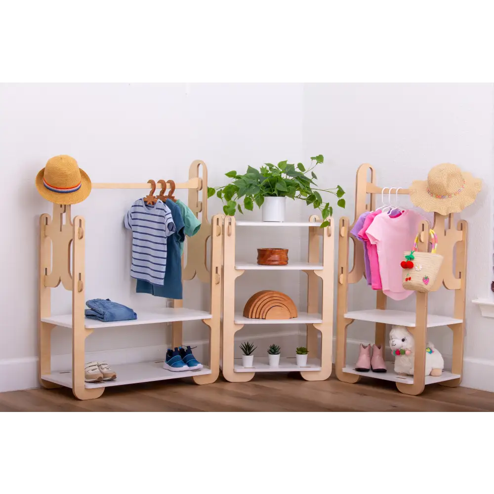 A charming display of children's wooden furniture set against a white wall, featuring two clothes racks with a selection of small, neatly hung garments including a striped shirt and a pink tunic, each rack adorned with a straw hat. The central shelving unit is delightfully decorated with a lush green plant, a wooden rainbow toy, and small potted succulents, with shoes and a plush toy on the lower shelves, creating an organized and playful space.